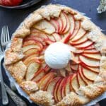 apple galette with a black background and ice cream on top with whole red apples also on the table