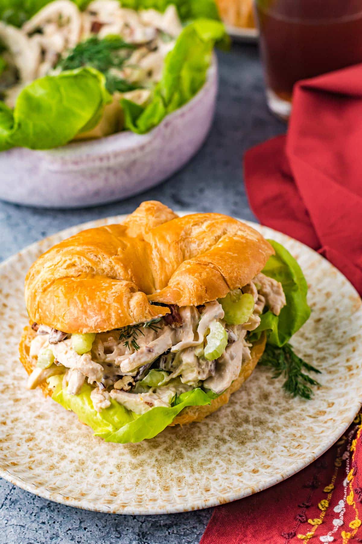 chicken salad sandwich on a croissant bun, filled to the max with celery and pecans, sitting on a speckled plate next to a red napkin