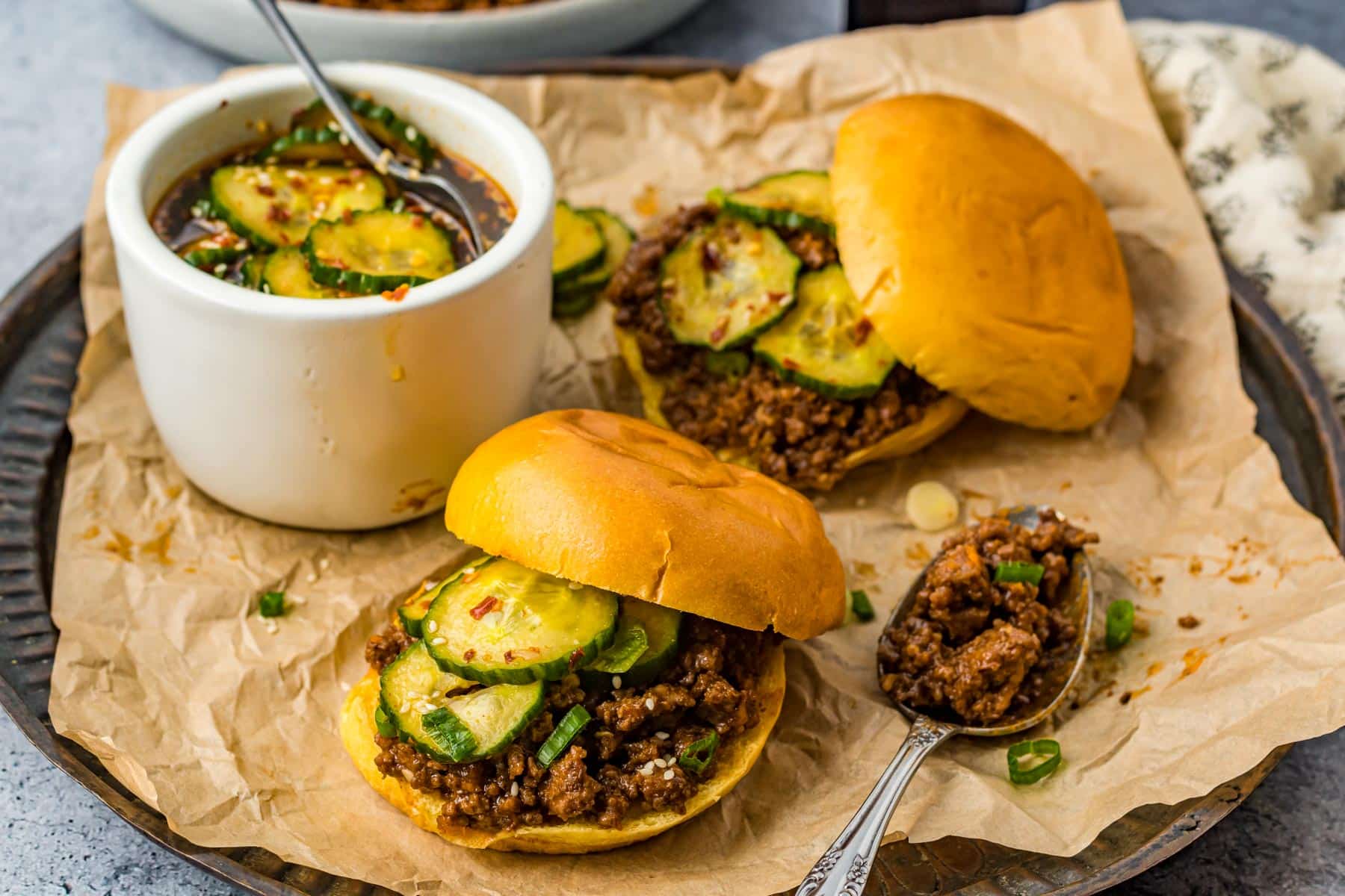 two sloppy Joe sandwiches on parchment paper with a bowl of quick pickles and a spoon full of beef mixture on a metal tray