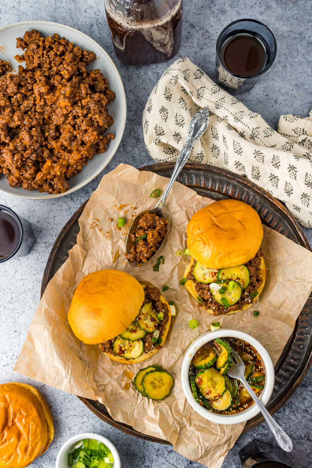 tray with ground beef bowl, bowl of pickles, and kitchen towel next to two sloppy joe sandwiches