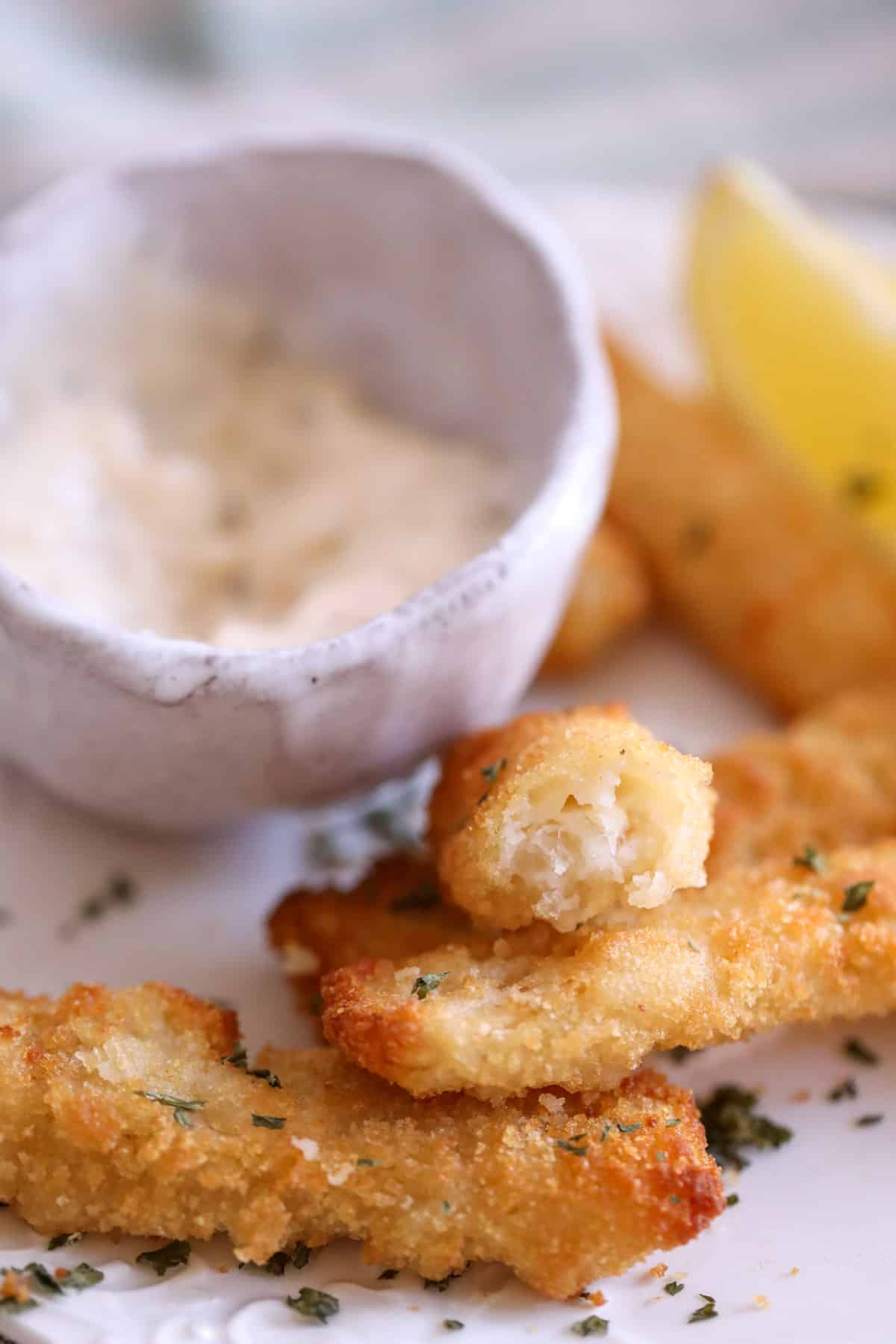 crunchy fish stick broken in half next to a side of tartar sauce with lemon wedges