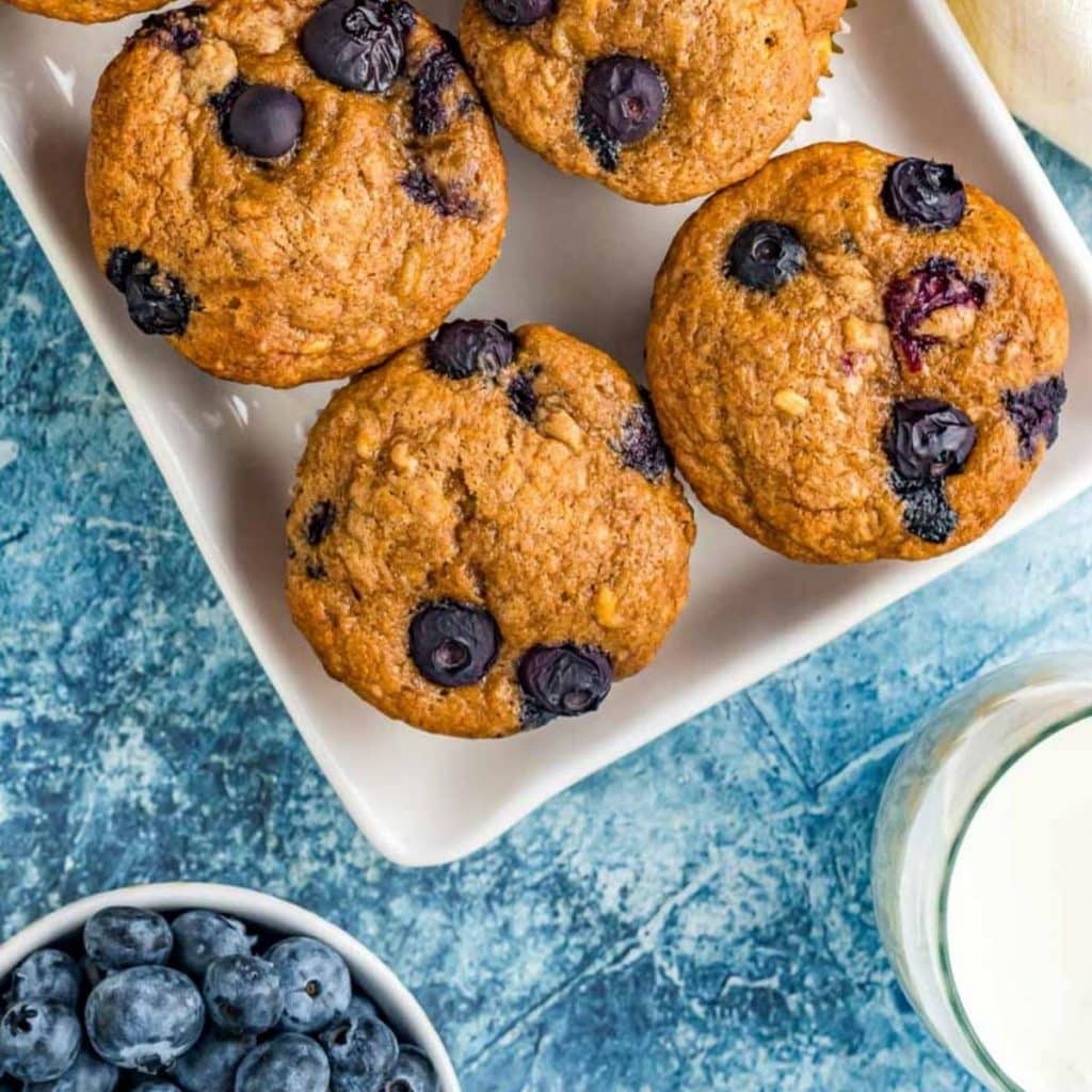 overhead view of four muffins with blueberries on top next to milk and fresh blueberries