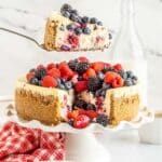 slice of berry cheesecake lifted from a cake stand and a red and white linen
