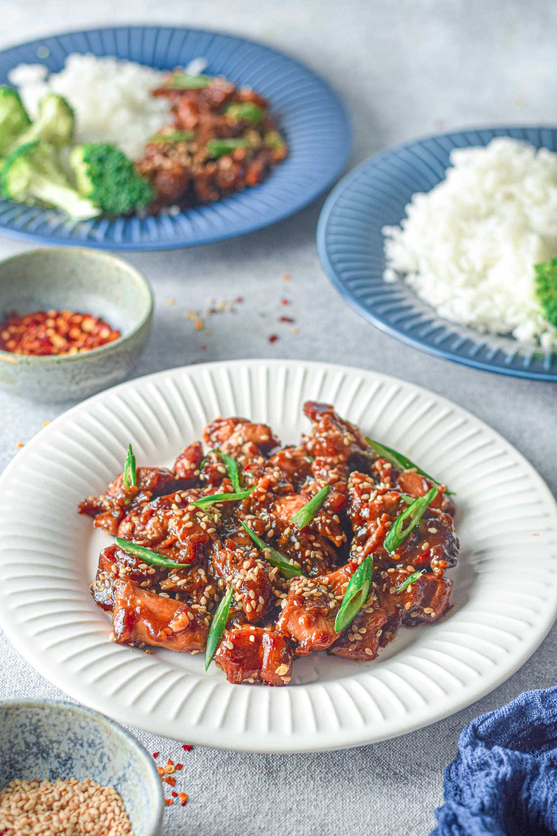 plate of teriyaki chicken with rice, broccoli next to a bowl of red pepper flakes and sesame seeds