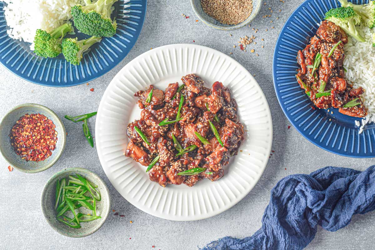 several plates on a table with teriyaki chicken garnished with green onion next to a bowl of red pepper flakes and a blue kitchen linen