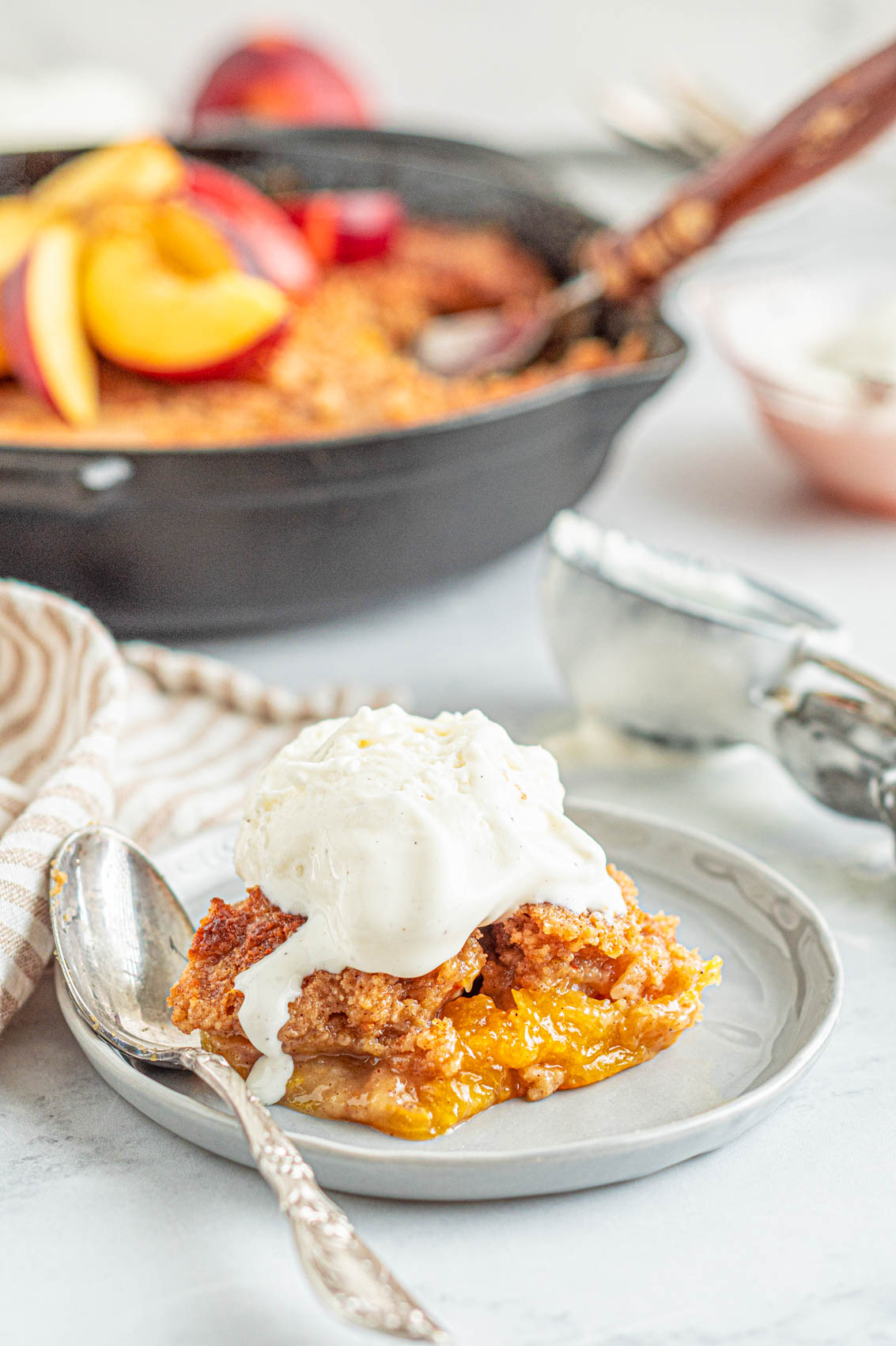 peach cobbler on a plate with a spoon and vanilla ice cream melting over the top with a cast iron skillet in background