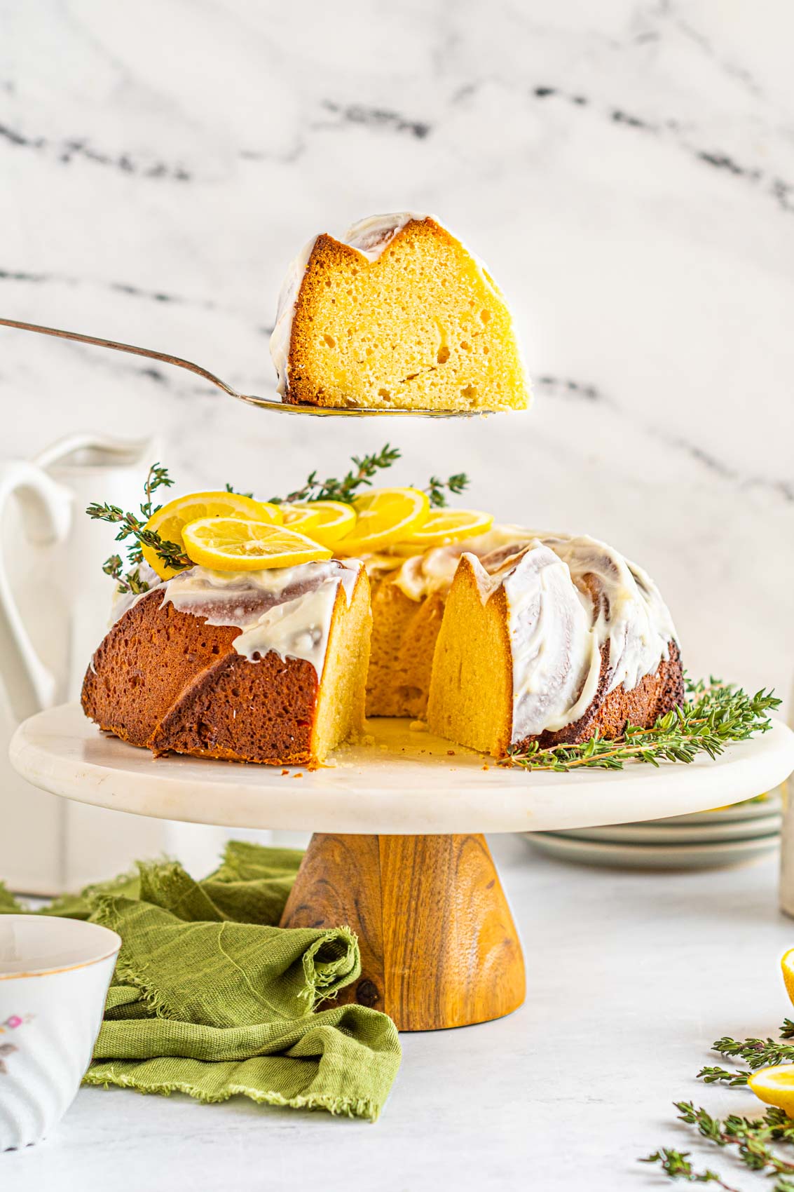 spatula lifting a piece of lemon bundt cake from the cake stand with a green towel on table