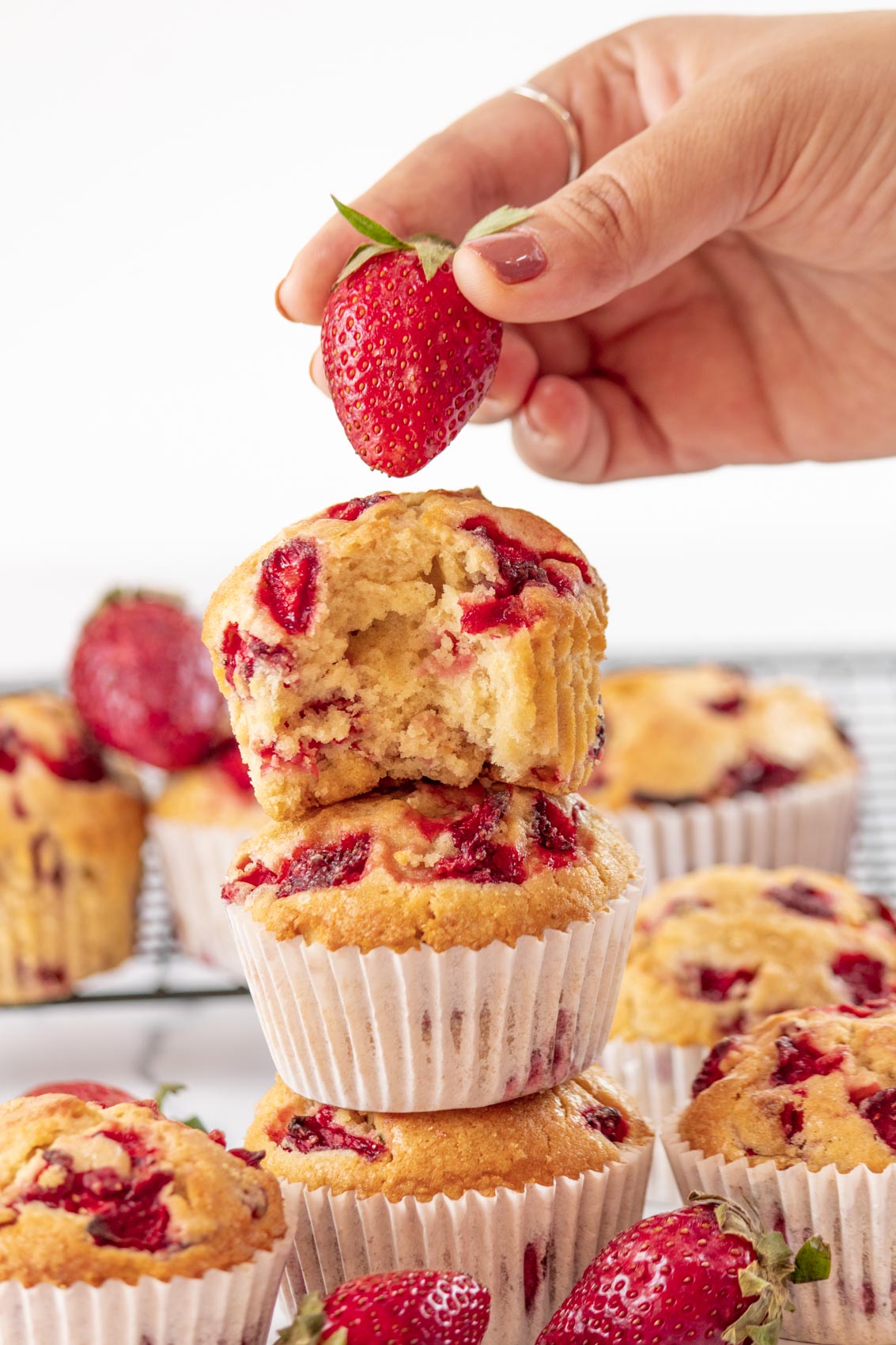 hand lifting a fresh strawberry next to a pile of strawberry muffins
