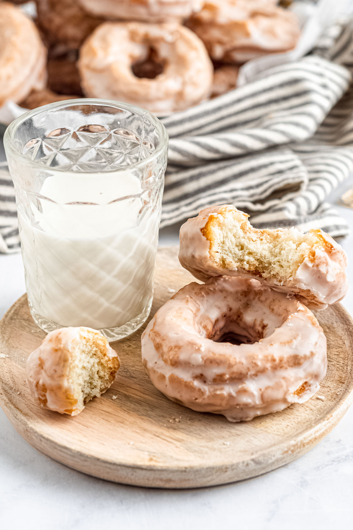 two glazed old-fashioned sour cream donuts with a glass of milk next to a striped linen; one donut has a large bite taken out