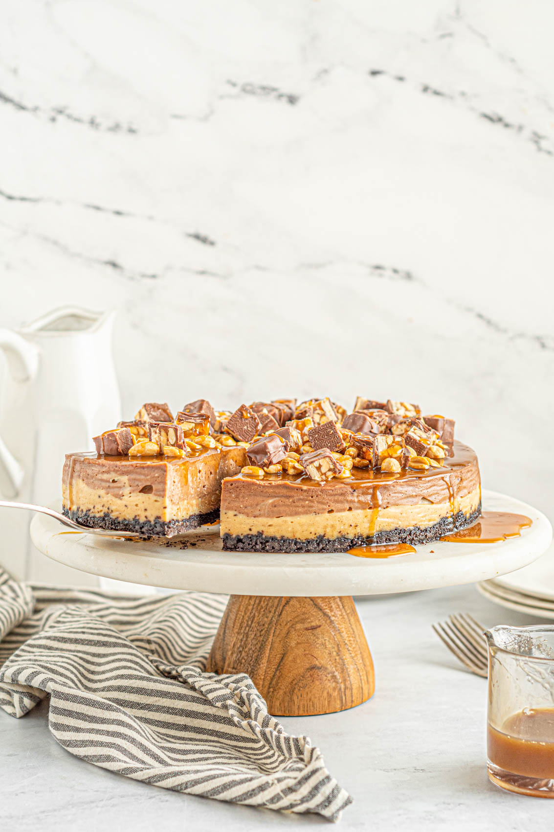 triple layer Snickers cheesecake on a cake stand next to a glass jar of caramel sauce